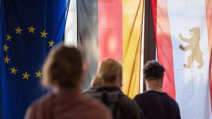 Archivbild: Wähler stehen in einem Wahllokal vor einer Europa-, Deutschland- und Berlinflagge (l-r). (Quelle: dpa/S. Gollnow)