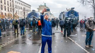 Ein Mann macht ein Selfie vor einer Demo in Berlin, im Hintergrund stehen Wasserwerfer (Quelle: dpa/Vladimir Menck/SULUPRESS.DE)
