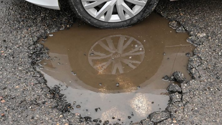 Symbolbild: Ein Pkw steht mit dem Vorderrad in einem mit Regenwasser gefüllten Schlagloch auf einer Straße. (Quelle: dpa/Pleul)