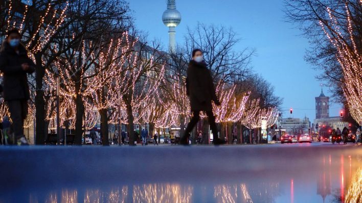 Passanten mit Mund-Nasen-Bedeckung überqueren die weihnachtlich beleuchtete Straße Unter den Linden.
