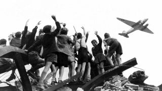 West-Berliner Jungen, die auf einem Trümmerberg stehen, begrüßen winkend ein US-amerikanisches Transportflugzeug, das Versorgungsgüter nach West-Berlin bringt (Bild: dpa, Archivfoto von 1948).