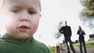 Symbolbild: Ein Junge mit laufender Nase blickt in die Kamera. Im Hintergrund sind seine Eltern auf einem herbstlichen Landweg zu erkennen. (Quelle: imago images/U. Grabowsky)