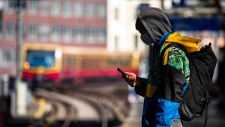 Symbolbild: Ein Jugendlicher mit Mundschutz blickt an einem Berliner S-Bahnhof auf sein Handy. (Quelle: imago images)