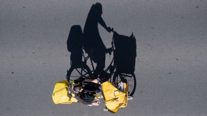 Ein Postbote auf einem Fahrrad wirft einen langen Schatten auf eine Straße (Quelle: dpa/Julian Stratenschulte)