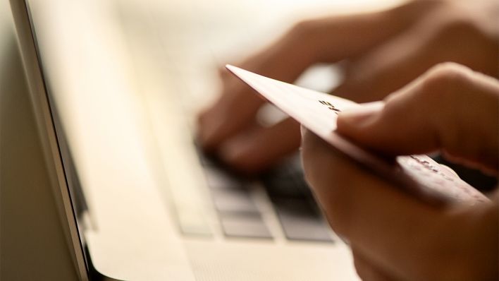 Symbolbild: Eine Person hält eine Kreditkarte in der Hand, während sie am Laptop sitzt (Bild: dpa/Franziska Gabbert)