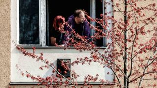 Eine Frau und ein Mann schauen in Berlin aus dem Fenster. Vor dem Fenster blüht ein Baum. (Quelle: dpa/Binh Truong)