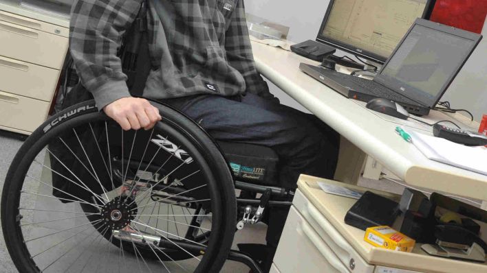 Symbolbild: Ein Mann sitzt in einem Rollstuhl an seinem Arbeitsplatz am Schreibtisch. (Quelle: dpa)