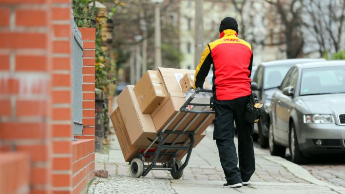 Symbolbild: Ein DHL Paketzusteller geht mit einer Sackkarre voll beladen mit Paketen zu einem Haus (Quelle: dpa/Jan Woitas)