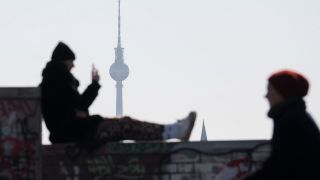 Ein Mann genießt das schöne Wetter und sitzt auf einer Mauer und schaut auf sein Smartphone, im Hintergrund steht der Fernsehturm. (Quelle: dpa/Jörg Carstensen)