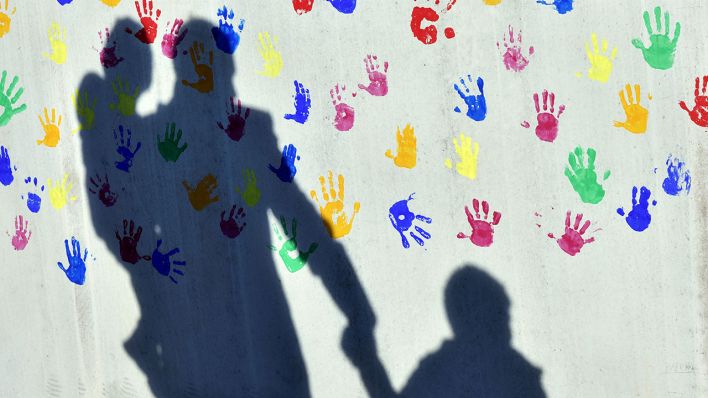 Ein Mann mit einem Kind auf dem Arm und einem an der Hand wirft einen Schatten auf eine mit bunten Handabdrücken bemalte Wand, Symbolbild (Quelle: DPA/Peter Kneffel)