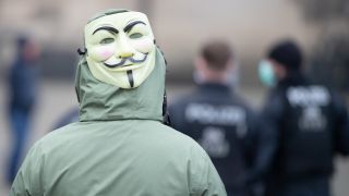 Eine Frau mit einer Guy-Fawkes-Maske steht hinter Polizisten. (Quelle: dpa/Sebastian Kahnert)