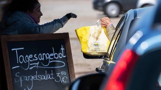 Eine Mitarbeiterin gibt am Drive-In-Schalter an einem kleinen Verkaufsstand auf dem Parkplatz einem Autofahrer eine Papiertüte mit warmem Essen (Bild: dpa/Soeren Stache)