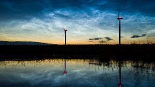 In einer Pfütze spiegeln sich die Landschaft und Windräder mit sogenannten leuchtenden Nachtwolken am 05.07.2016 kurz vor Mitternacht über Sieversdorf (Brandenburg). (Quelle: dpa/Patrick Pleul)