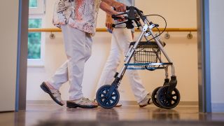 Symbolbild: Eine ältere Frau geht mit einer Gehhilfe, in Begleitung einer Pflegekraft über den Flur einer Pflegeeinrichtung. (Quelle: dpa/C. Schmidt)