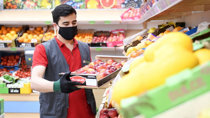Ein Mitarbeiter eines Lebensmittelgeschäfts füllt in der Gemüseabteilung ein Regal auf und trägt eine Maske als Mundschutz. (Quelle: dpa/Tobias Hase)