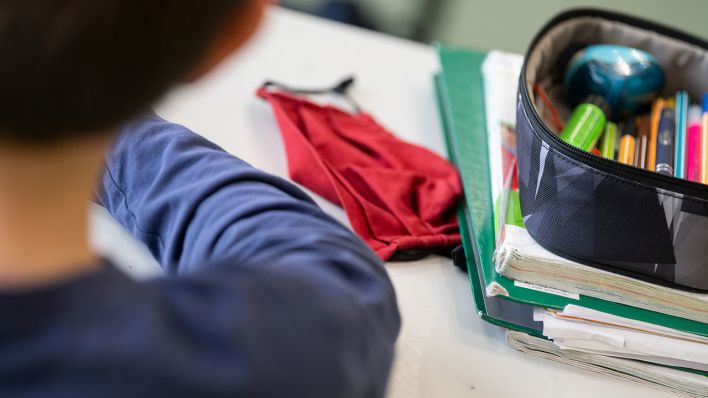 Eine Mund-Nasen-Bedeckung liegt während einer Unterrichtsstunde neben einem Mäppchen und Schulbüchern auf einem Schultisch. (Quelle: dpa/Marijan Murat)