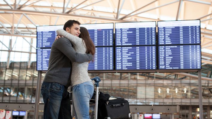 Ein Mann und eine Frau am Flughafen vor einem Display mit den Ankunfts- und Abflugszeiten und umarmen sich. (Quelle: dpa/Christin Klose)