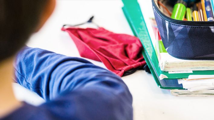 Symbolbild: Eine Mund-Nasen-Bedeckung liegt während einer Unterrichtsstunde einer fünften Klasse neben einem Mäppchen und Schulbüchern auf einem Schultisch. (Quelle: dpa/Marijan Murat)