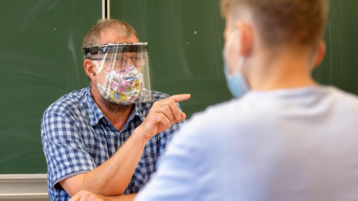 Symbolbild: Ein Lehrer mit Mundschutz und Gesichts-Schild sitzt vor SchülerInnen in einem Klassenzimmer. (Quelle: imago images/R. Unkel)