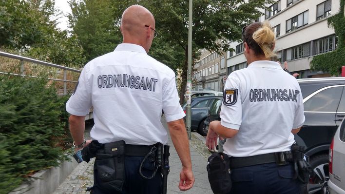 Zwei Mitarbeiter des Ordnungsamtes tragen weiße Hemden, auf denen "Ordnungsamt" steht, während sie am 27.08.2019 durch eine Straße in Kreuzberg gehen. (Quelle: dpa/Wolfram Steinberg)