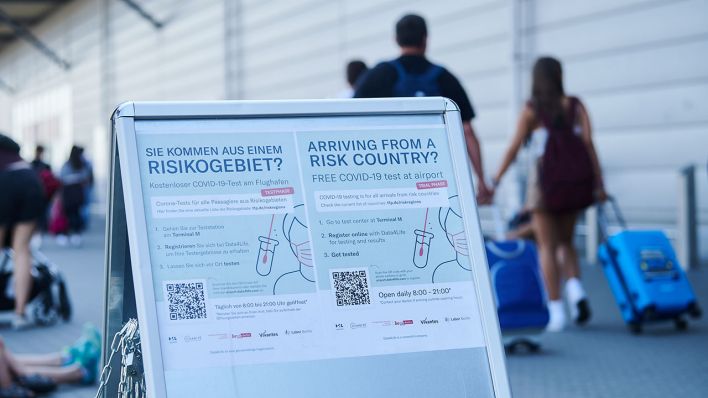 "Sie kommen aus einem Risikogebiet?" und weitere Informationen stehen auf einem Schild am Flughafen Schönefeld, während Passagiere mit Rollkoffern vorbeilaufen (Quelle: dpa/Annette Riedl)