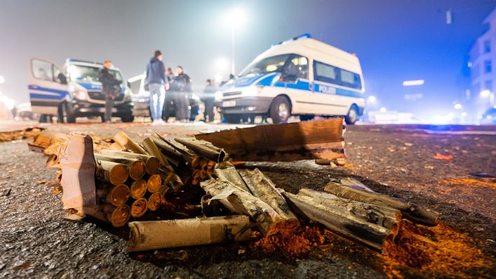 Symbolbild: Gesprengte Böller liegen auf der Straße, im Hintergrund sind Polizei-Einsatzkräfte und -Fahrzeuge zu sehen. (Quelle: dpa/A. Arnold)