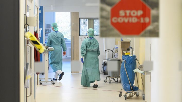 Symbolbild: Intensivpfleger laufen in der Corona-Intensivstation eines Krankenhauses über den Gang während im Vordergrund ein Schild mit der Aufschrift "Stop Covid-19" an der Tür zu sehen ist. (Quelle: dpa/Robert Michael)