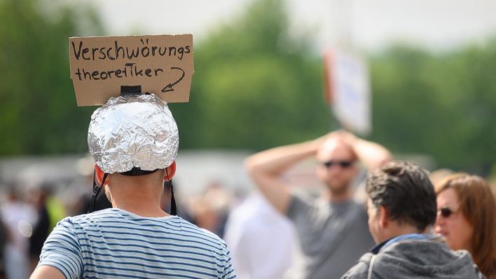 Ein Mann trägt während einer "Querdenken"-Kundgebung einen Aluhut mit einem Schild, auf dem "Verschwörungstheoretiker" steht (Quelle: dpa/Sebastian Gollnow)