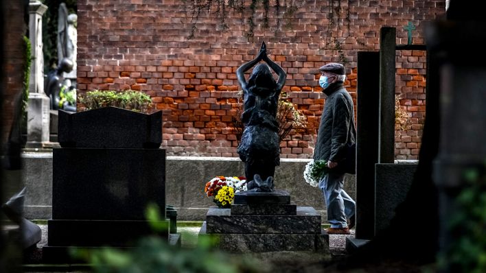 Symbolbild: Ein Mann mit Mund-Nase-Bedeckung geht auf einem Friedhof an Gräbern vorbei und trägt einen Blumenstrauß. (Quelle: dpa/C. Furlan)