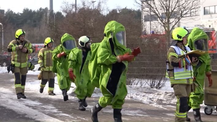 Mitarbeiter der Katastrophenschutzes laufen auf den Hof eines Recycling-Unternehemens, auf dem 1.000 Liter Salzsäure ausgeflaufen sind, sie tragen grüne Schutzanzüge. (Quelle: TNN)