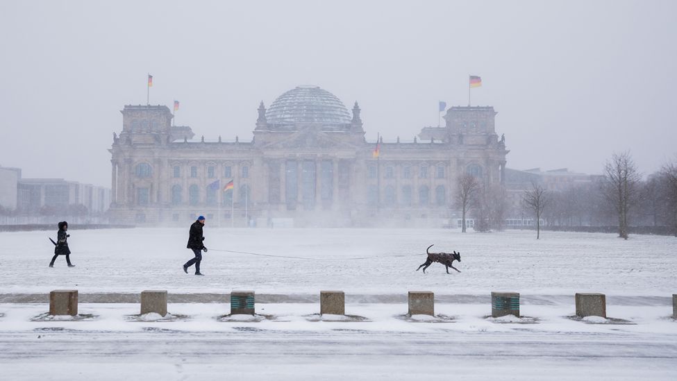 Spaziergänger mit Hund gehen am 07.02.2021 bei Schneefall vor dem Reichstagsgebäude. (Quelle: dpa/Christoph Soeder)