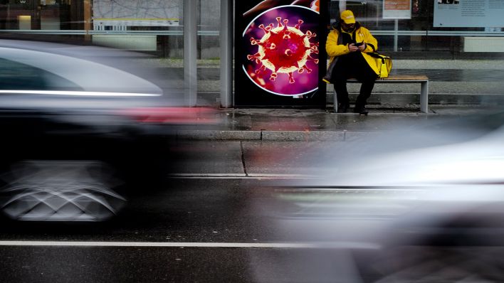 Symbolbild: Ein Mann wartet an einer Bushaltestelle neben einem Plakat mit einem Modell eines Coronavirus'. (Bild: dpa/Markus Schreiber)
