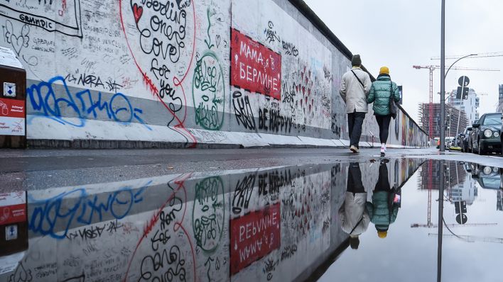 Bei Regen gehen am 06.01.2021 zwei Personen an der Berliner Mauer entlang und spiegeln sich in einer Pfütze. (Quelle: dpa/Kira Hofmann)