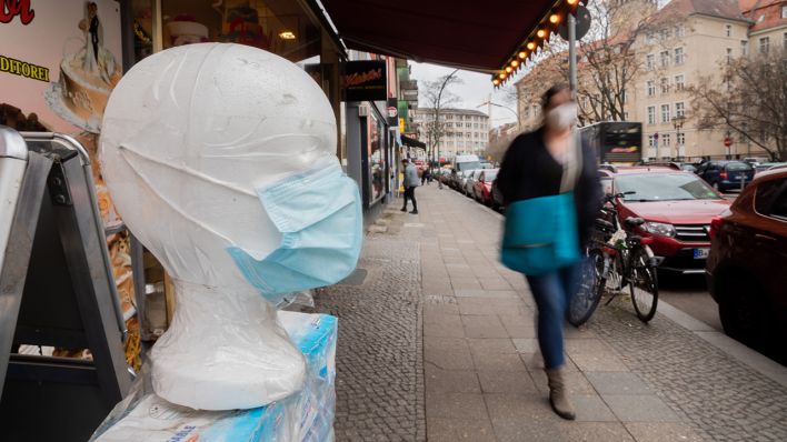 Eine medizinische Maske wird vor einem Geschäft in Berlin-Neukölln ausgestellt, während eine Passantin vorbeigeht. (Quelle: dpa/Christoph Soeder)