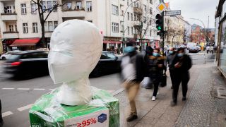 Eine FFP2-Maske wird vor einem Geschäft in Berlin-Neukölln ausgestellt, an dem Passanten vorbeigehen (Quelle: dpa/Christoph Soeder)
