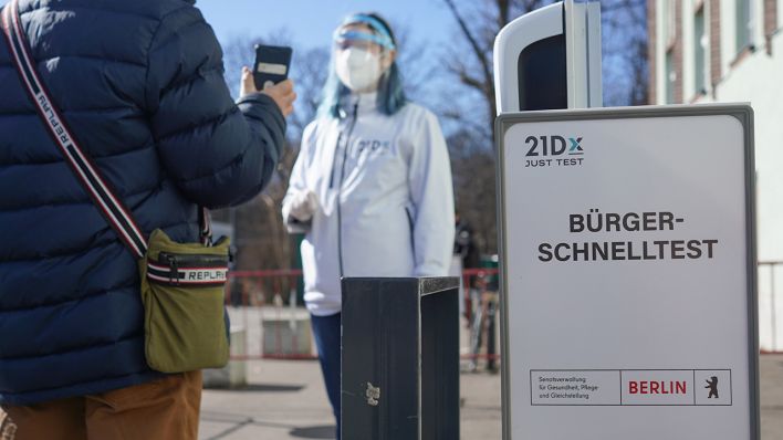 Symbolbild: Eine Mitarbeiterin der Betreiber-Firma 21Dx GmbH und ein Bürger stehen vor einem Corona-Schnelltestzentrum in der Lehrter Straße hinter einem Schild mit der Aufschrift «Bürger-Schnelltest». (Quelle: dpa/J. Carstensen)