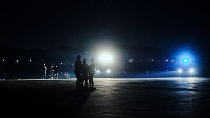 Scheinwerfer eines Polizeifahrzeugs beleuchten am späten Abend Personen, die sich auf dem Tempelhofer Feld aufhalten. (Quelle: dpa/Stefan Jaitner)