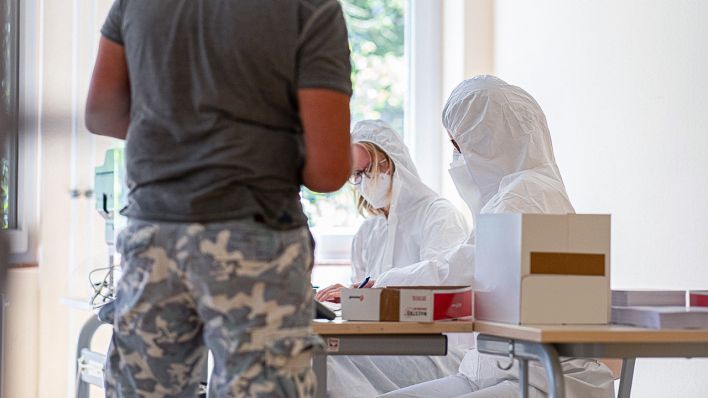 Symbolbild: Zwei Mitarbeiterinnen im Schutzanzug nehmen bei einem Mann beim gratis Coronavirus-Test am Carl-Miele-Berufskolleg die Daten auf. (Quelle: dpa/Guido Kirchner)
