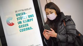 Junge Frau mit Mundschutz,MaskeHinter ihr eine Leuchtreklame fuer die Corona Warn App. Sie blickt auf Ihr Smartphone. (Quelle: dpa/Sven Simon)