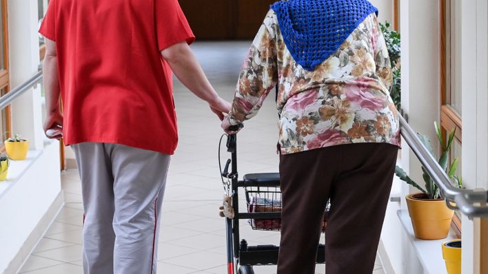 Ein Mann und eine Seniorin mit einem Rollator gehen durch den Flur eines Pflegeheims. (Quelle: dpa/Jens Kalaene)