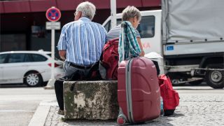 Eine Frau und ein Mann sitzen in Berlin mit Koffern vor einem Flughafen. (Quelle: dpa/Paul Zinken)