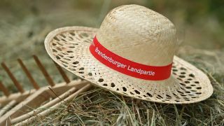 Ein Strohhut mit der Aufschrift "Brandenburger Landpartie" liegt auf einer Harke und einem Heuhaufen in einem Garten. (Quelle: dpa/Patrick Pleul)