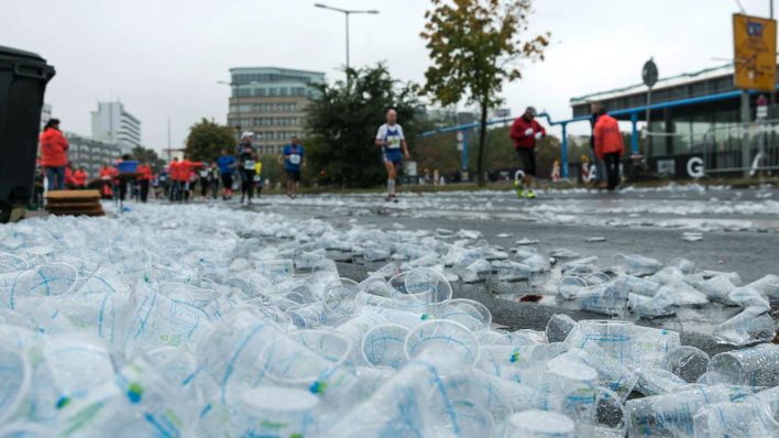 Ein Haufen leerer Plastikbecher liegt auf der Straße, im Hintergrund laufen Marathonläufer. Bild: imago-images/POP-EYE
