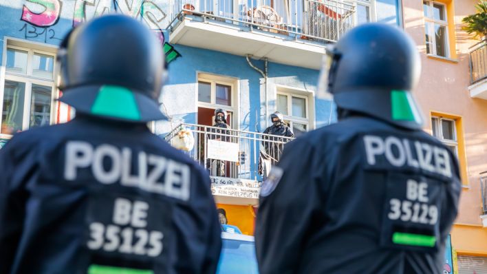 Polizisten stehen bei einem Polizeieinsatz in der Rigaerstraße 94 im Berliner Bezirk Friedrichshain vor dem Gebäude des linken Hausprojekts, wobei weitere Beamte auf einem Balkon stehen. (Quelle: dpa/Christoph Soeder)
