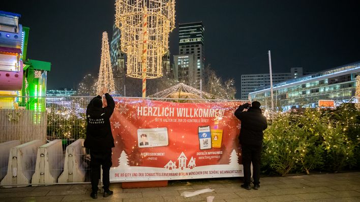 Symbolbild: Zwei Sicherheitsmitarbeiter hängen am Weihnachtsmarkt auf dem Breitscheidplatz ein Hinweisbanner zum 2G-Zutritt auf. (Quelle: dpa/C. Soeder)