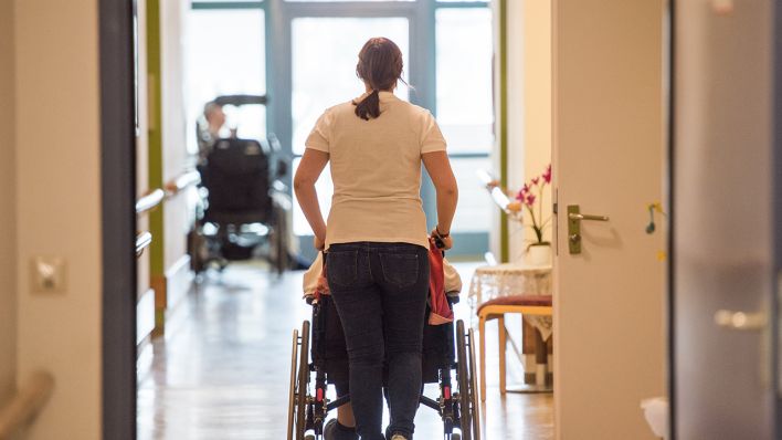 Eine Pflegerin schiebt eine ältere Frau im Rollstuhl durch einen Flur. (Quelle: dpa/Christophe Gateau)