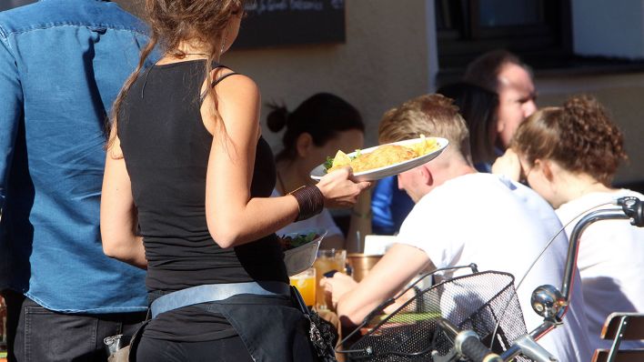 Eine Kellnerin trägt Speisen und Getränke an einem Tisch im Außenbereich eines Cafés. (Quelle: imago-images/Ralph Peters)