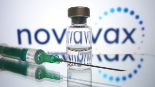 Symbolbild: Vor einem Schild mit der Aufschrift "Novavax" liegt eine Phiole und eine Spritze (Bild: dpa/Sven Simon)