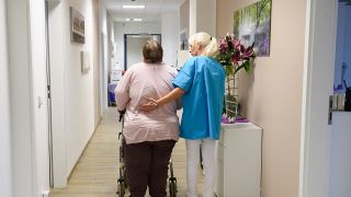 Eine Pflegerin läuft mit einer Patientin über den Flur einer Pflegeeinrichtung (Bild: dpa/Hans Wiedl)