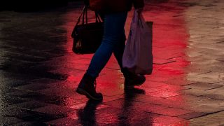 Eine Frau mit einer Einkaufstüte geht am verkaufsoffenen Sonntag durch die Innenstadt auf einem Gehweg, auf den rotes Licht aus einem Schaufenster fällt (Quelle: dpa / Daniel Bockwoldt) .